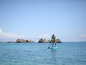 Žena stojící na paddleboardu v moři s kameny v pozadí.