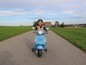 Dívka jede na modrém skútru Vespa po štěrkové cestě.