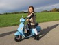 Dívka jede na modré elektrické motorce, podobné Vespe.