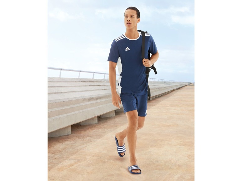 Muž v tričku a šortkách Adidas jde po betonové cestě v žabkách.
