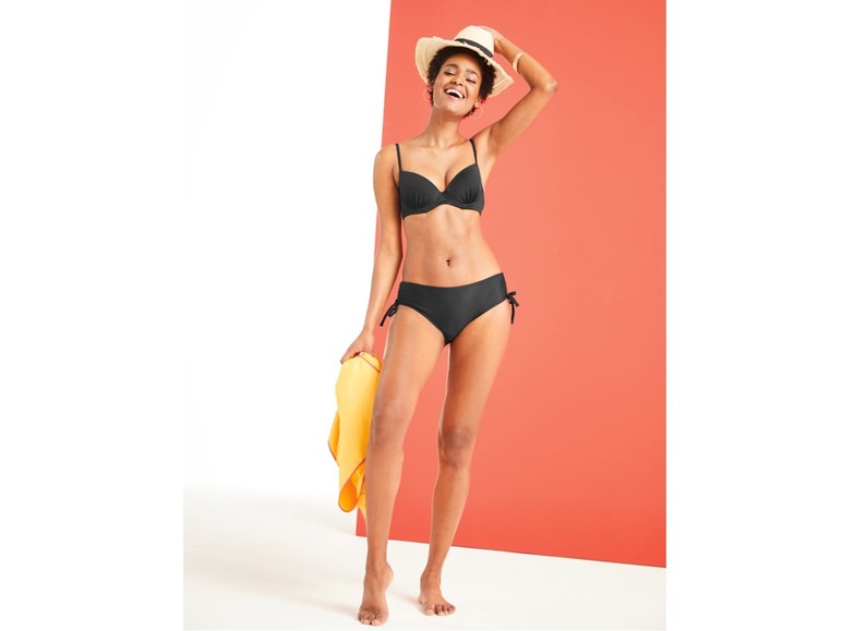 Žena v černých plavkách a slaměném klobouku s žlutým ručníkem v ruce.