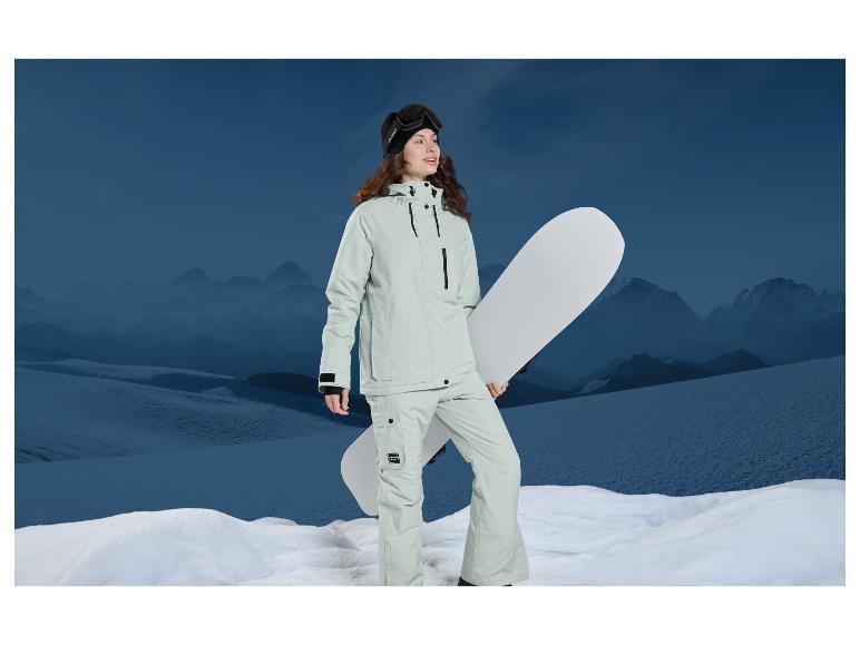 Žena v zeleném lyžařském overalu drží snowboard před zasněženými horami.