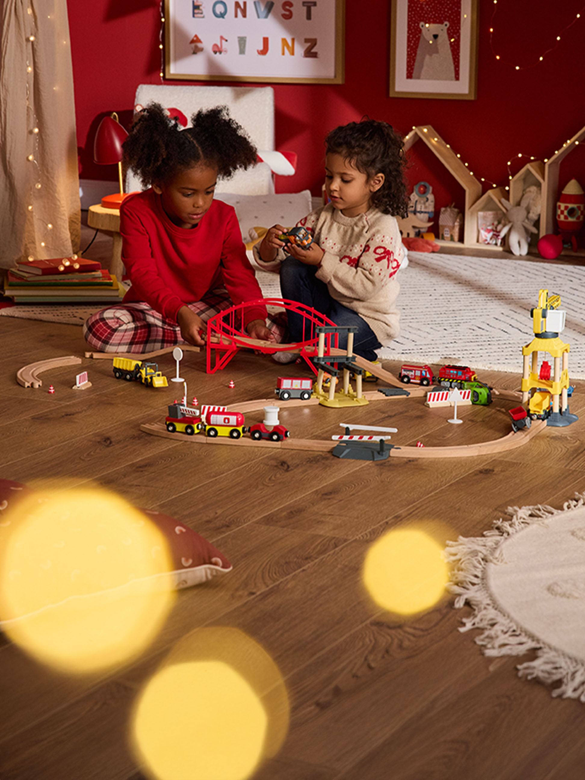 Dvě děti si hrají s dřevěnou vláčkodráhou a vozidly na dřevěné podlaze.