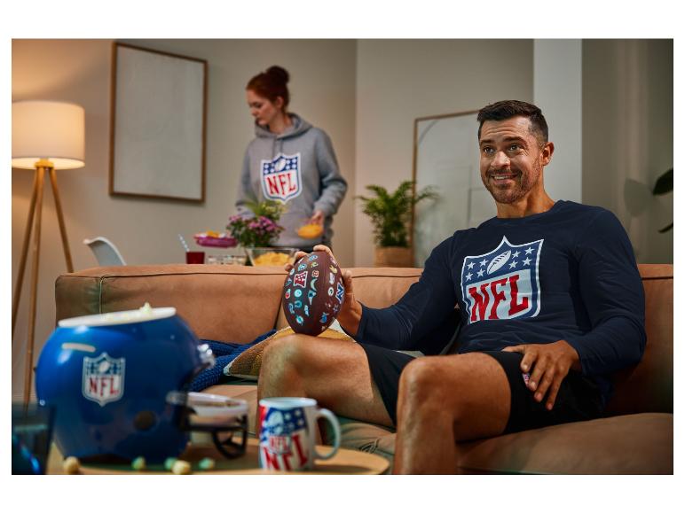 Muž v tričku s logem NFL drží fotbalový míč na pohovce.