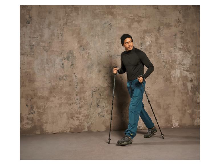 Muž s trekingovými holemi a trekingovými botami kráčí před betonovou stěnou.