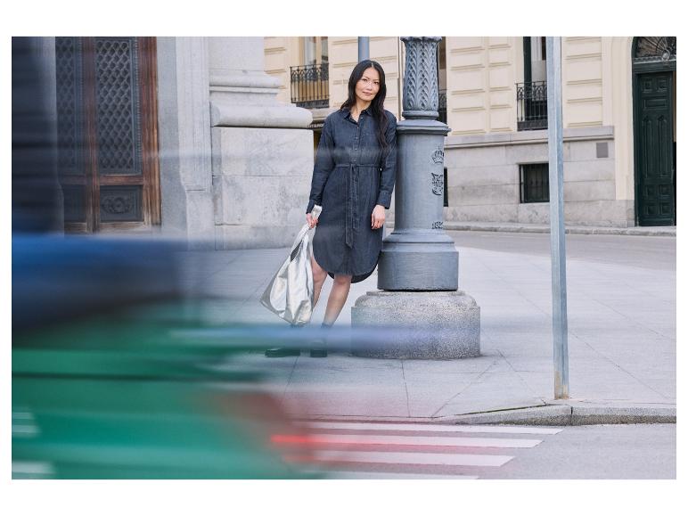 Žena v džínových šatech stojí u lampy a drží stříbrnou tašku.