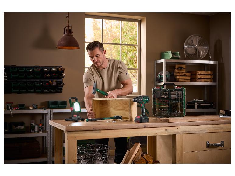 Muž sestavuje dřevěnou krabici kladivem, obklopen elektrickým nářadím a dílenským vybavením.