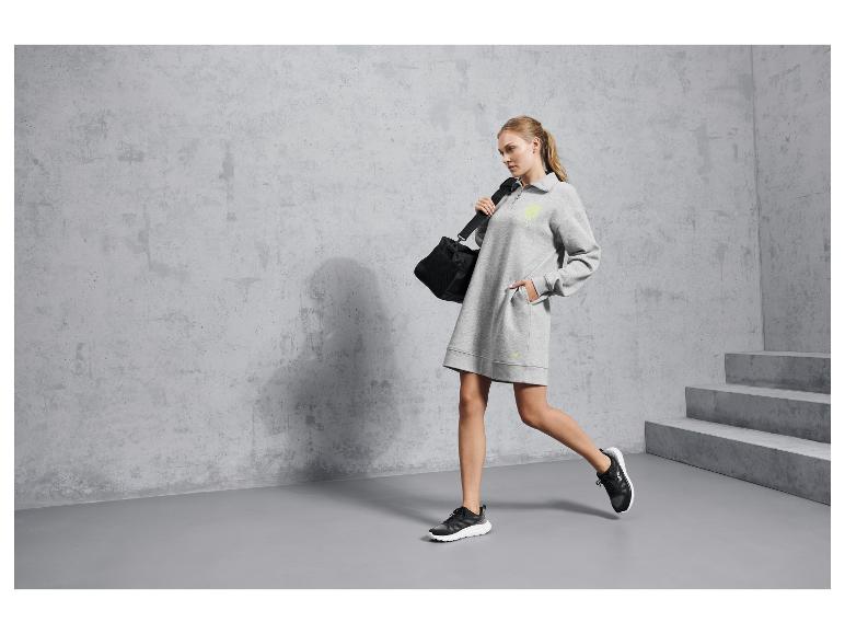 Žena v šedých sportovních šatech a černých Reebok botách, drží černou sportovní tašku.