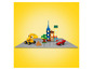 Žlutý LEGO bagr, červené auto a malé LEGO budovy na šedé podložce.