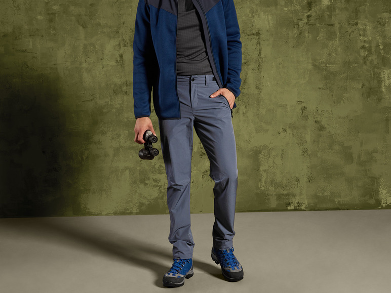 Muž v modré fleecové bundě, šedých kalhotách a modrých turistických botách.