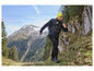 Muž v bundě a kalhotách s logem 'Exploring the Mountains' jde po horské stezce.