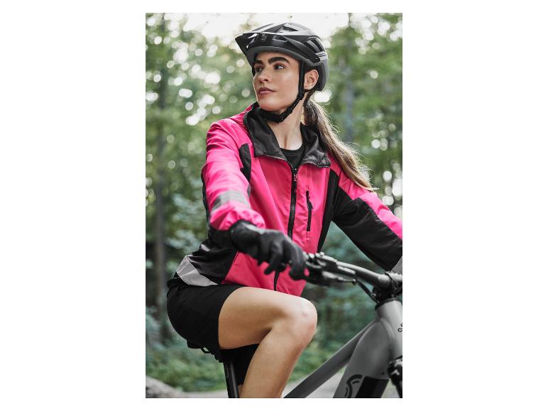 Žena v růžové cyklistické bundě a černých šortkách jede na horském kole.