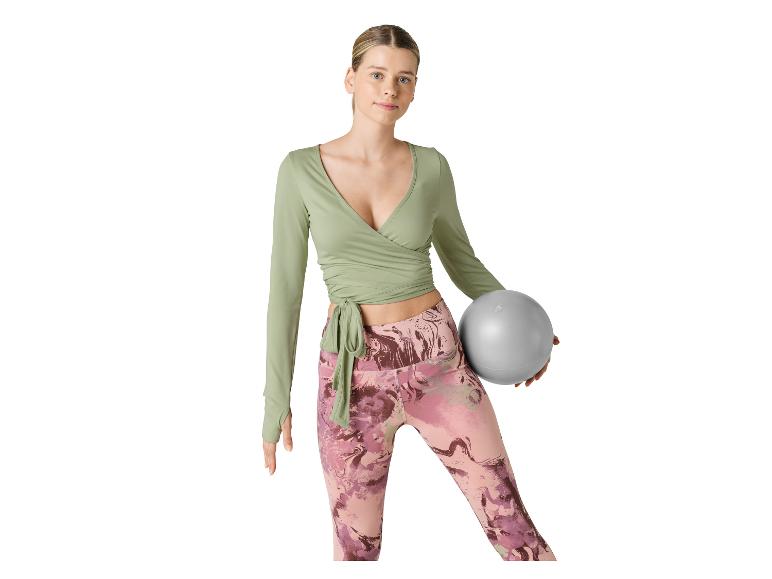 Žena v zeleném zavinovacím topu a vzorovaných legínách drží pilates míč.