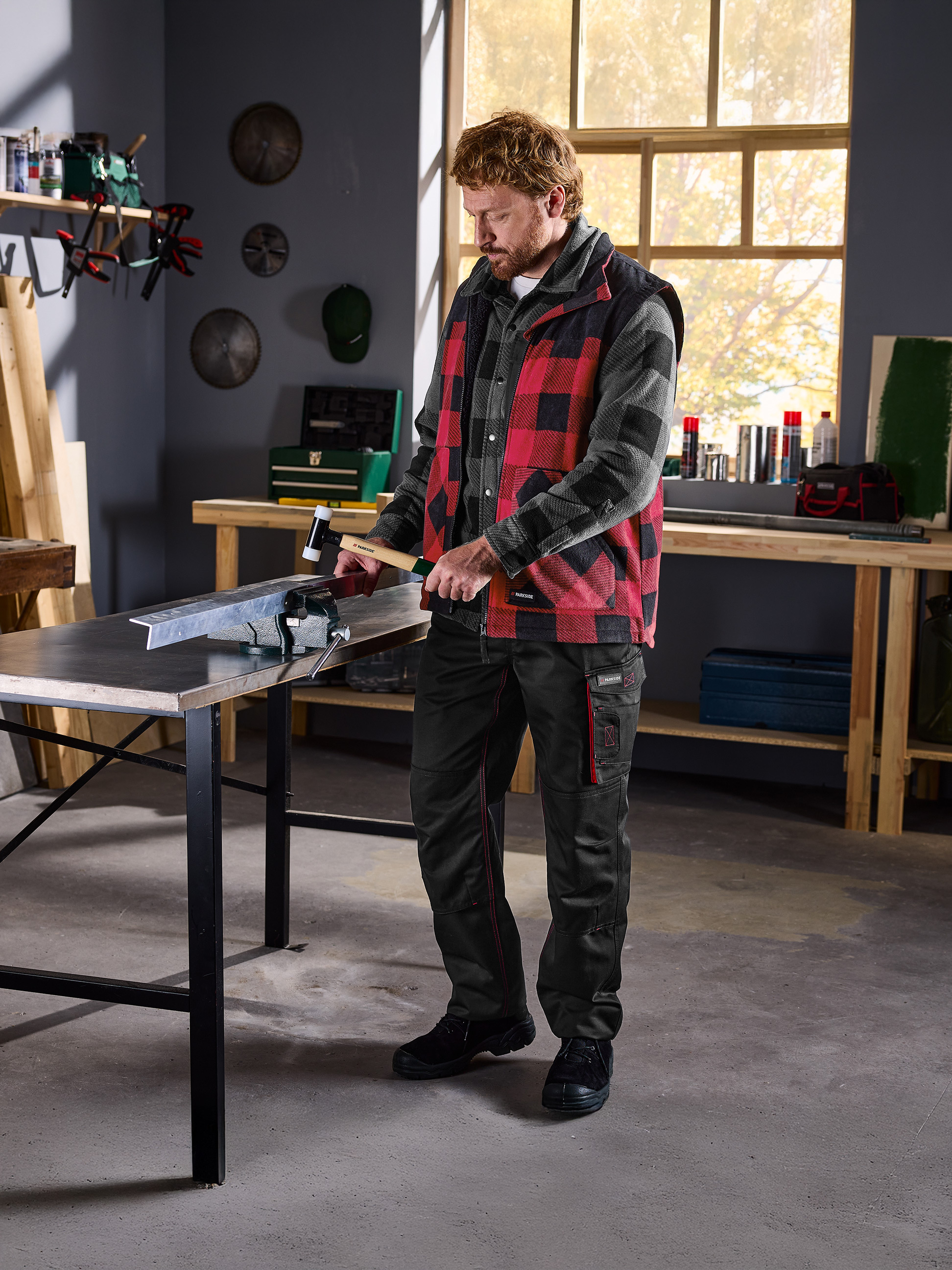 Muž v kostkované košili a vestě Parkside pracuje s kovem ve svěráku v dílně.