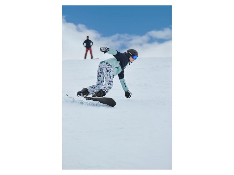 Snowboardista v vzorovaných kalhotách a světle modré bundě sjíždí zasněžený svah.