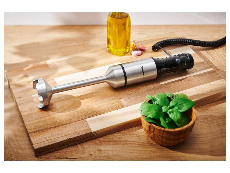 Tyčový mixér, láhev oleje, česnek a bazalka na dřevěném prkénku.