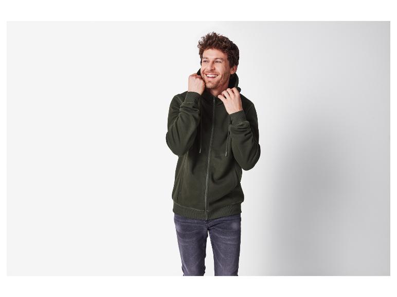 Usmívající se muž v tmavě zelené mikině na zip a šedých džínách.