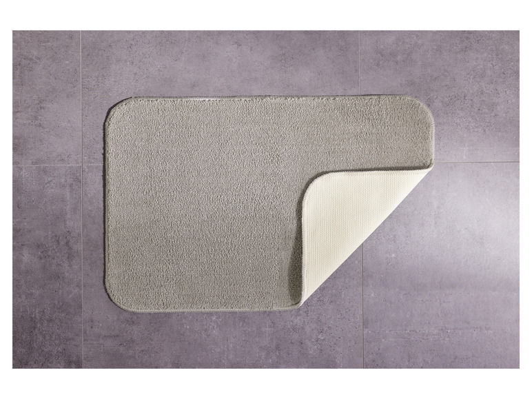 Šedý koupelnový koberec s béžovou spodní stranou na dlaždicové podlaze.