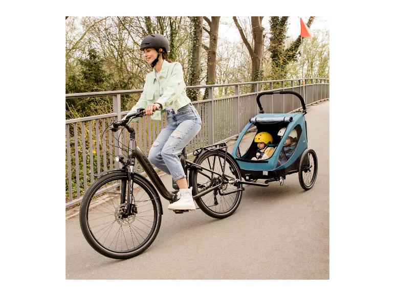 Žena na elektrokole táhne cyklistický vozík se dvěma dětmi.