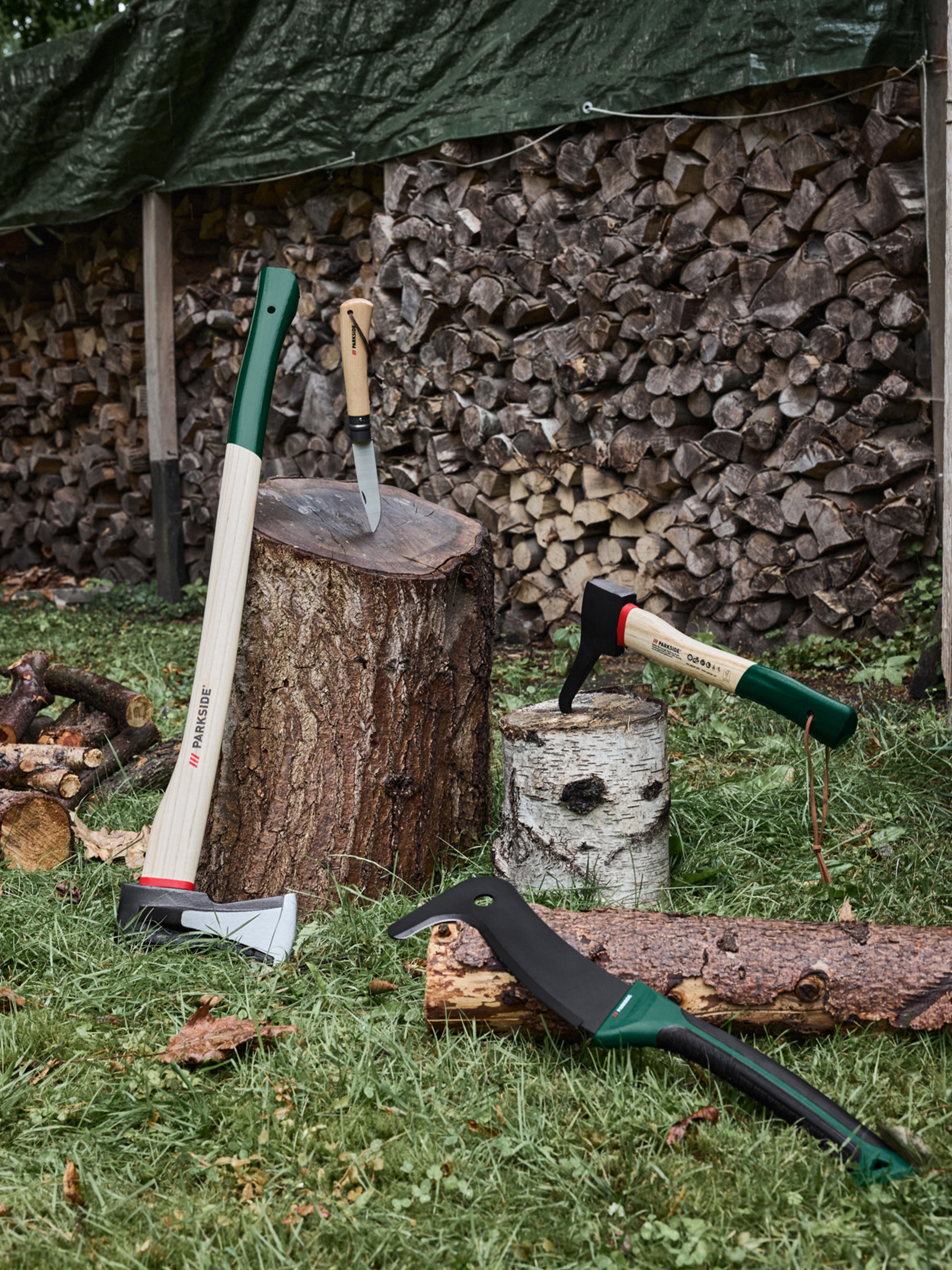 Sekery, nůž a mačeta na trávě s naskládaným dřevem v pozadí.