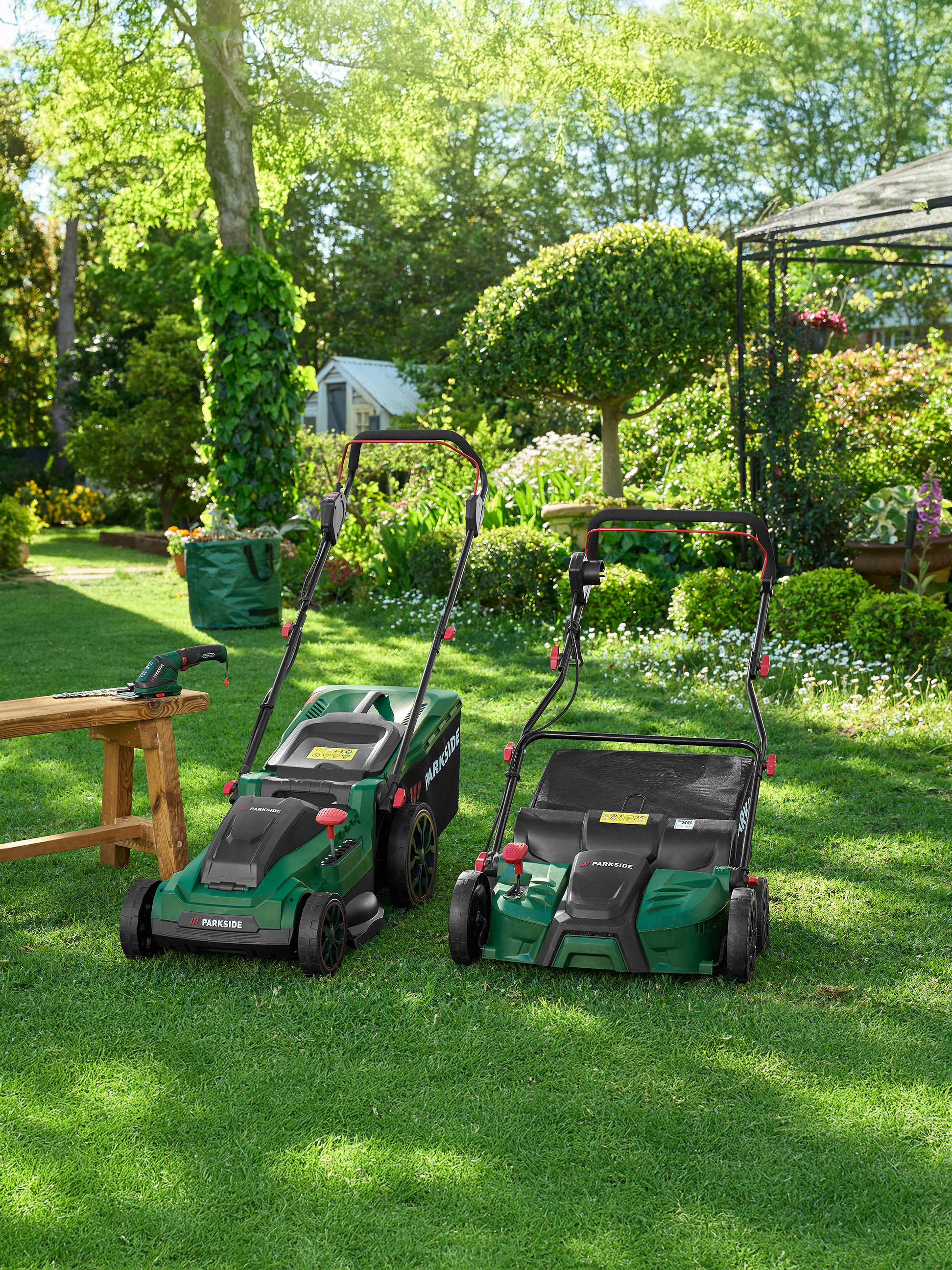 Dvě zelené sekačky Parkside a zahradní nářadí na trávníku v zahradě.