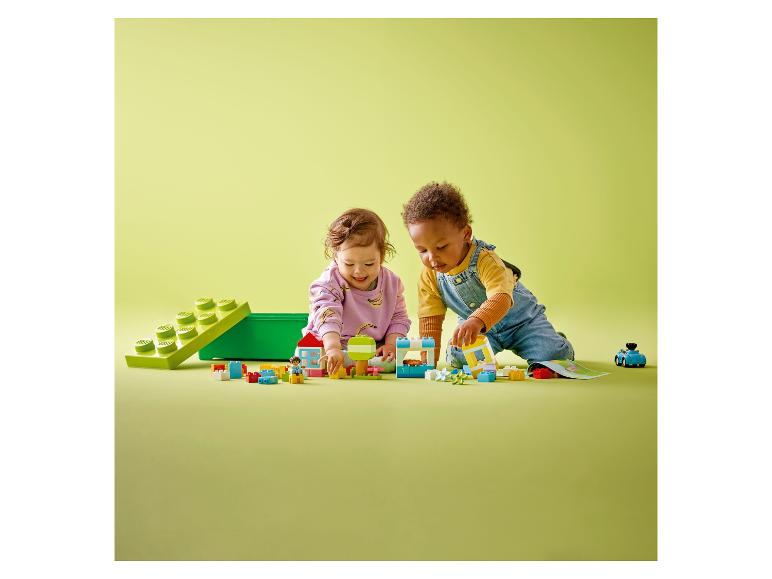 Dvě batolata si hrají s barevnými kostkami a figurkami LEGO DUPLO na zeleném povrchu.