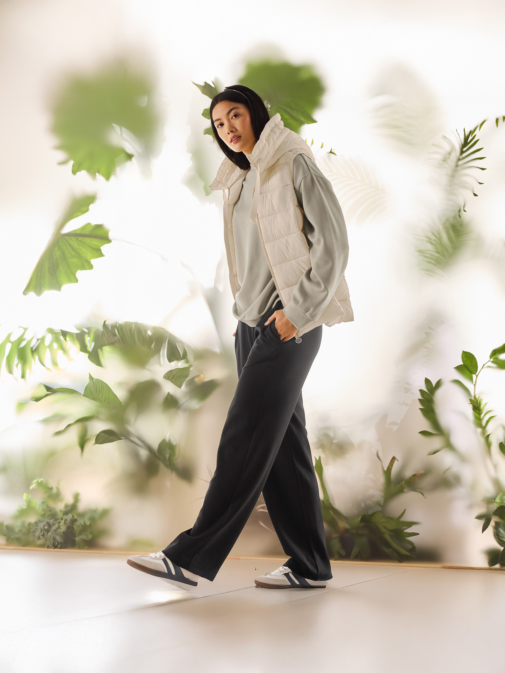 Žena v béžové prošívané vestě, světle zelené mikině a černých kalhotách kráčí.