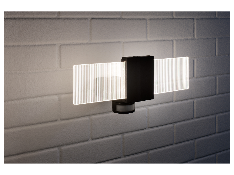 Moderní černá nástěnná lampa s pohybovým senzorem a LED osvětlením, namontovaná na bílé cihlové zdi.