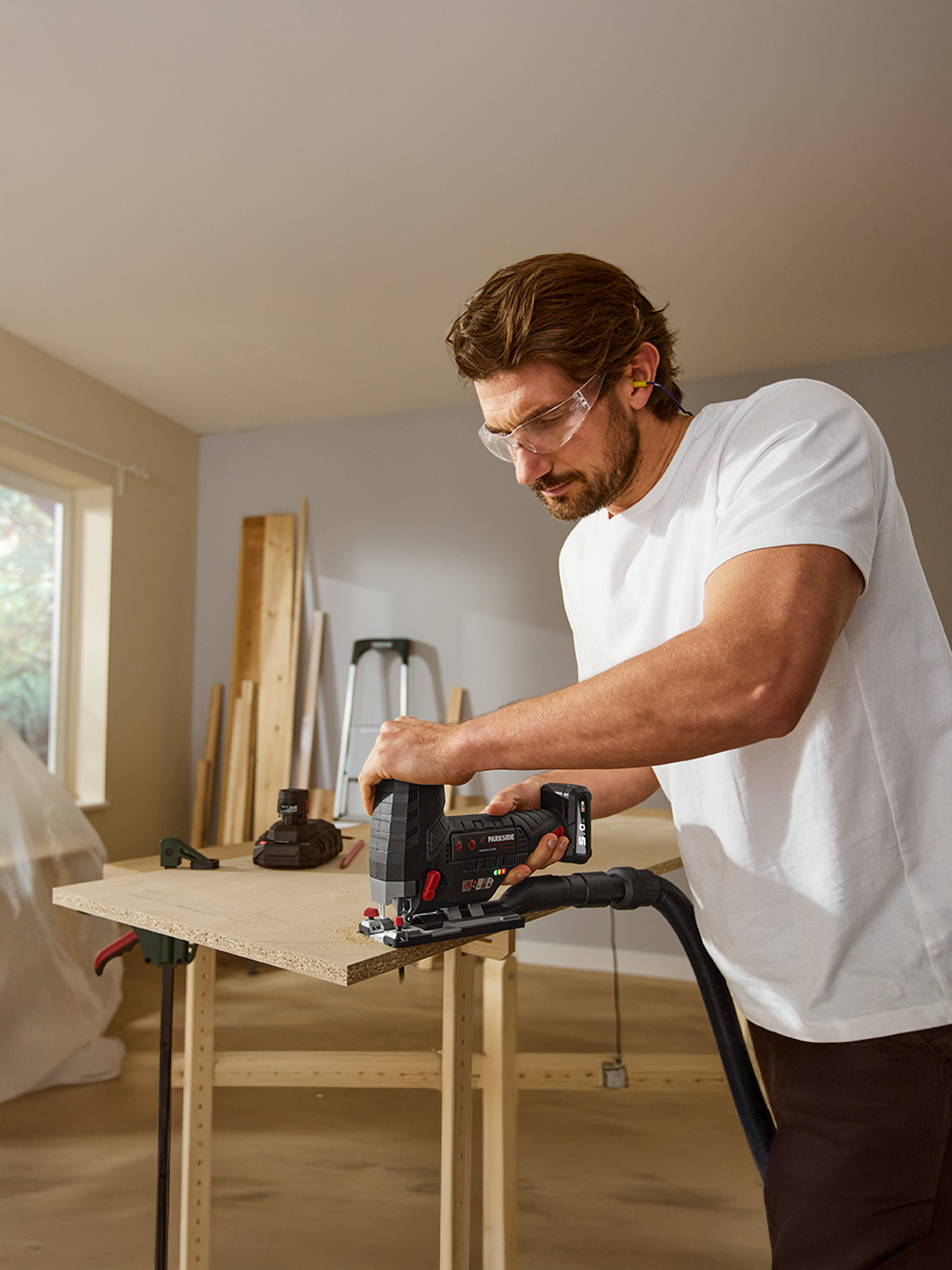Muž v ochranných brýlích a špuntech do uší řeže dřevo akumulátorovou přímočarou pilou.