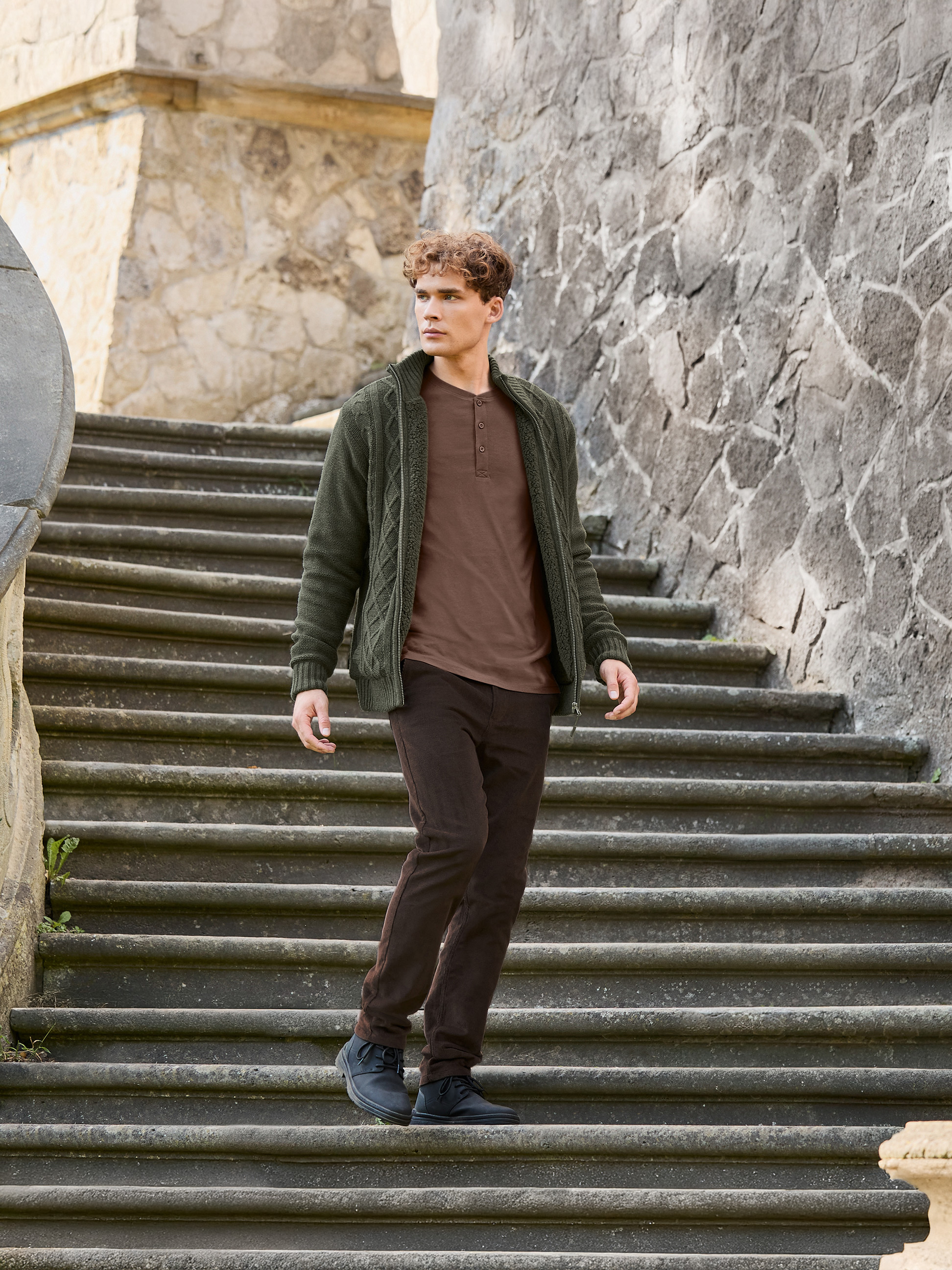 Muž v zeleném svetru, hnědém tričku a hnědých kalhotách schází po kamenných schodech.