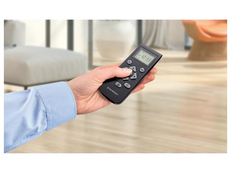 Ruka drží černý dálkový ovladač s digitálním displejem zobrazujícím 12:33, ve světlé místnosti.