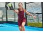 Žena v bordó sportovních šatech hraje padel s raketou a míčkem.