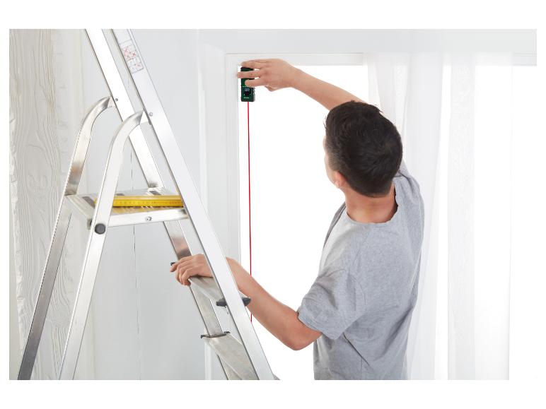 Muž měří okno laserovým dálkoměrem, zatímco stojí na žebříku.
