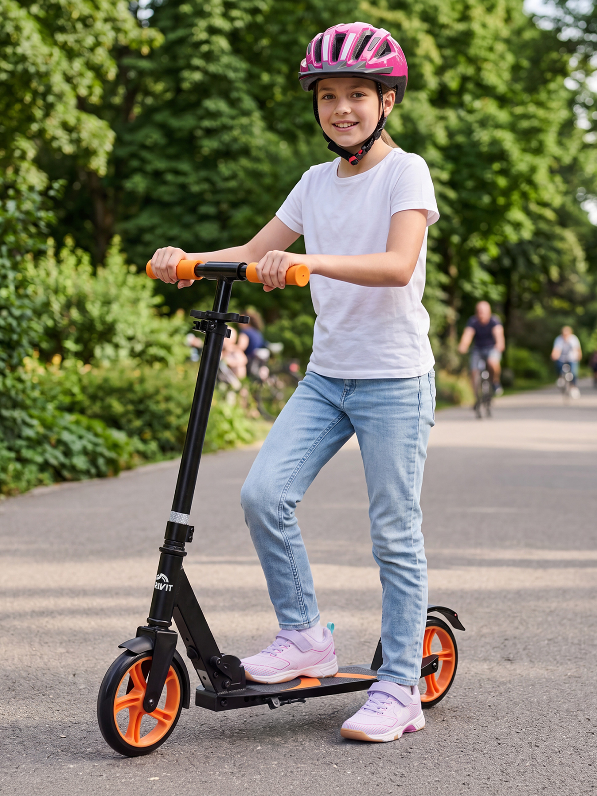 Dívka v růžové helmě a na černé koloběžce s oranžovými koly v parku.