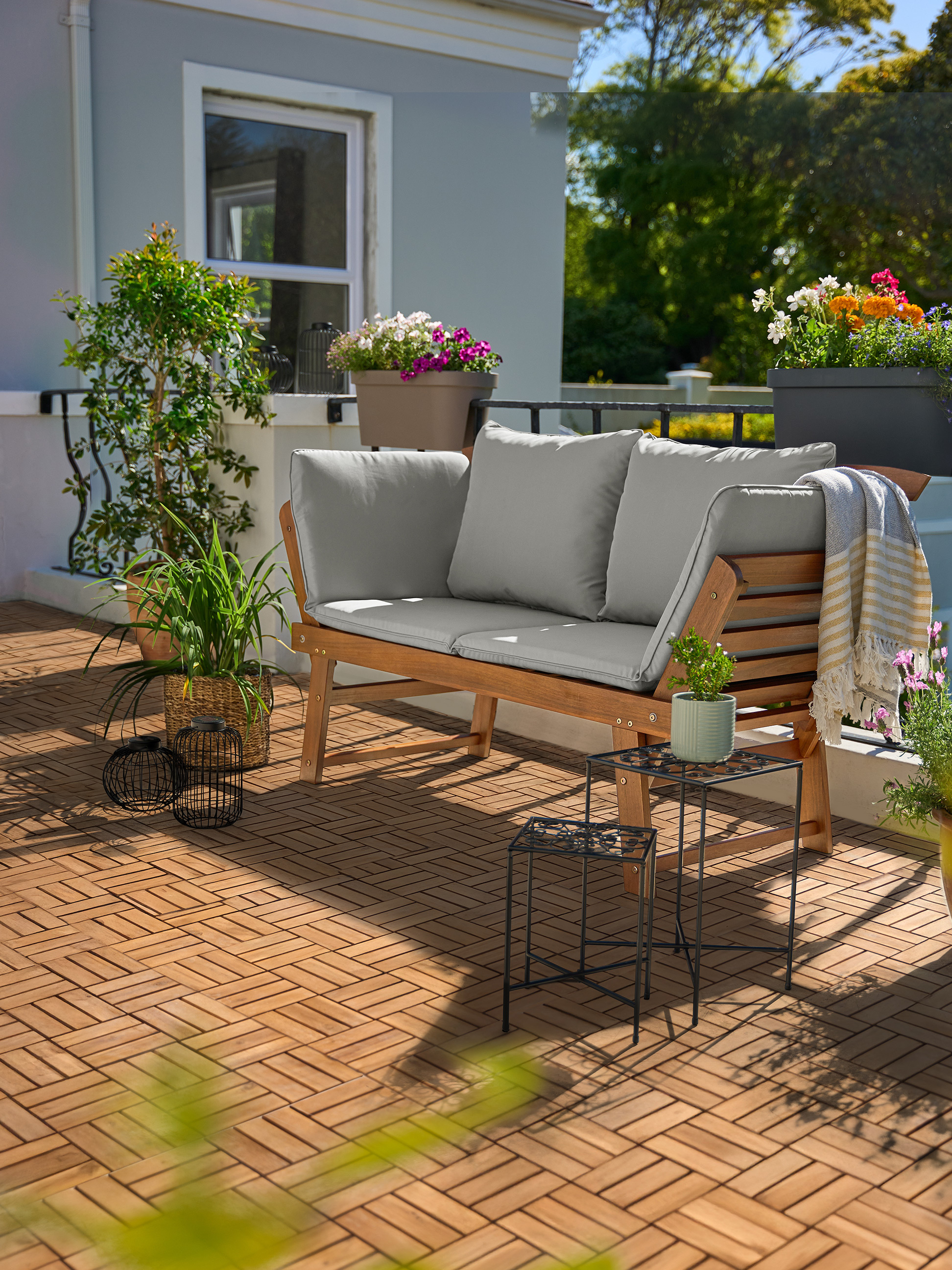 Dřevěná zahradní pohovka s šedými polštáři na balkoně s dřevěnými dlaždicemi a rostlinami