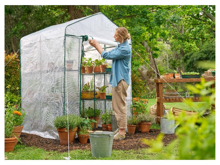 Žena otevírá malý skleník s rostlinami v květináčích a zahradním nářadím.