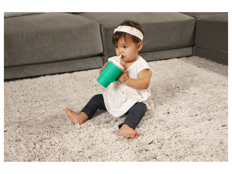 Dítě pije z hrnečku se slámkou na koberci.