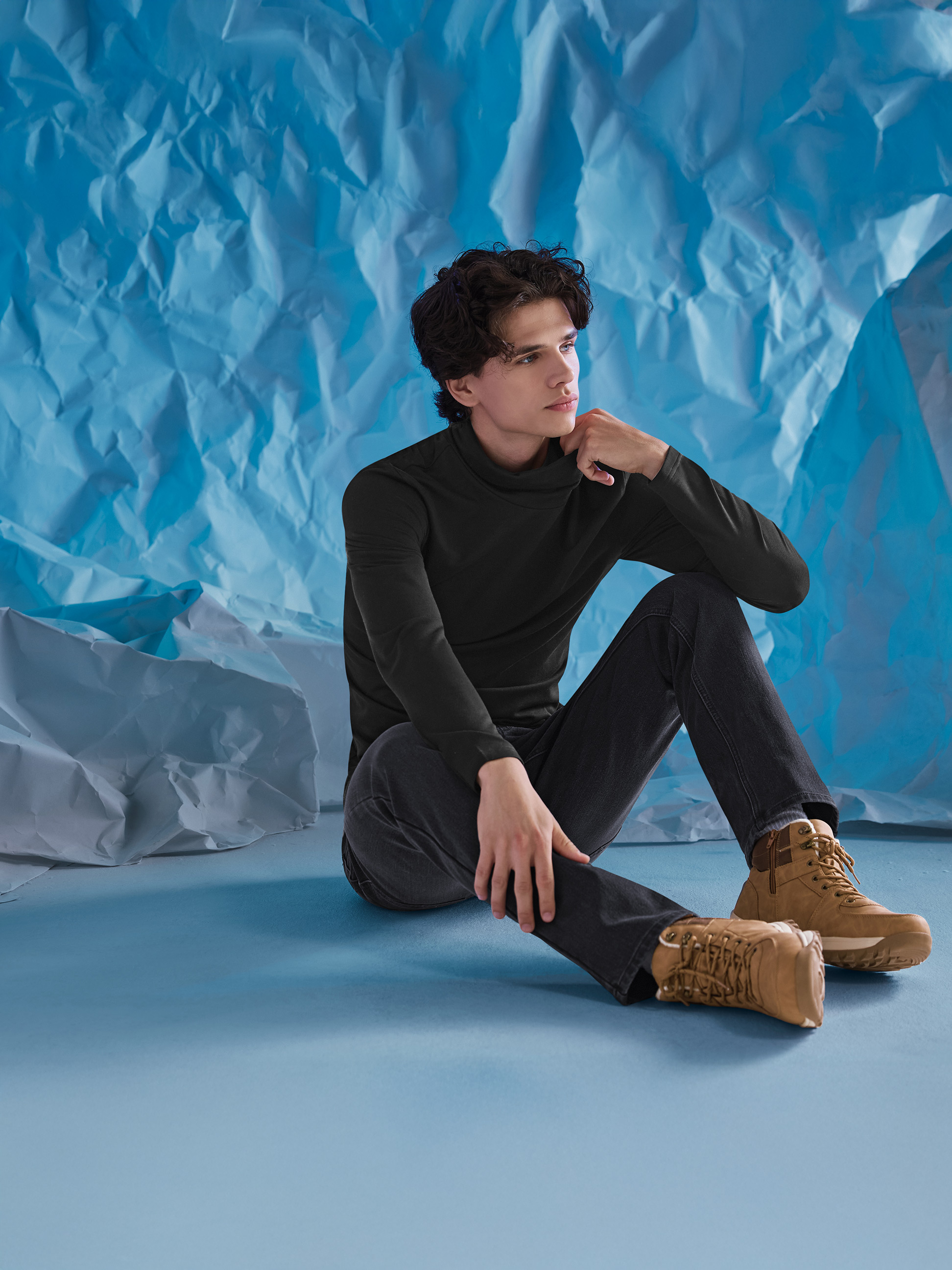 Muž v černém roláku, džínách a hnědých botách, sedící na modrém pozadí.