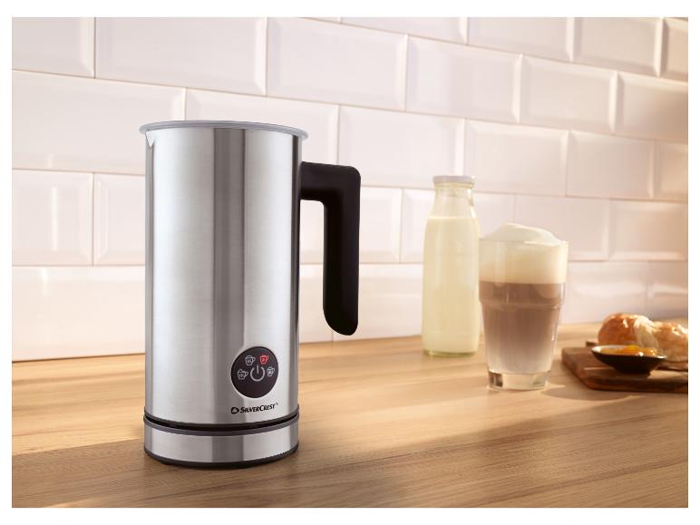 Nerezový napěňovač mléka na dřevěné desce se sklenicí latte a pečivem.