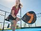 Žena hraje padel s černou a oranžovou padelovou raketou na modrém kurtu.