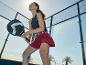 Žena ve sportovním oblečení s pálkou na padel na kurtu, usmívající se a vzhlížející.
