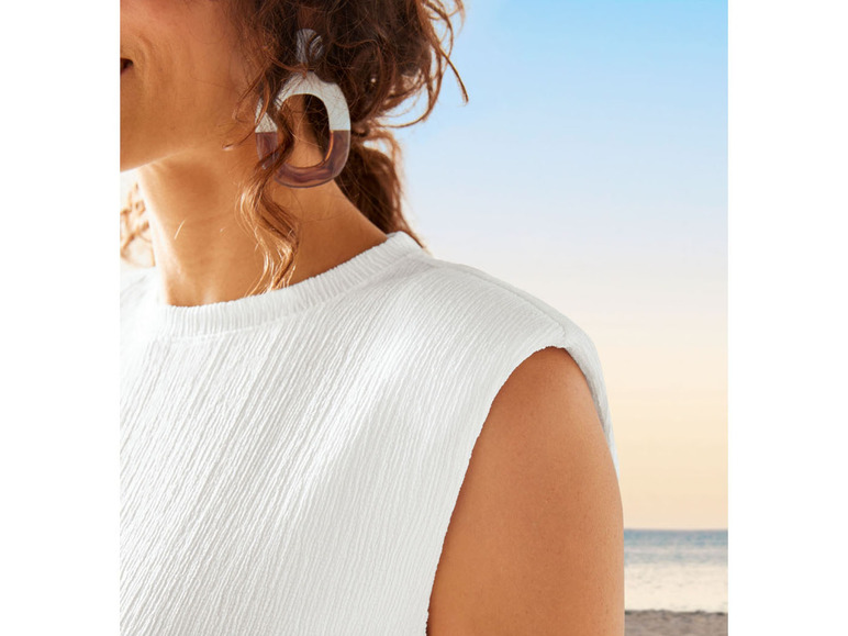 Žena v bílém topu s kapucí na pozadí pláže a moře.