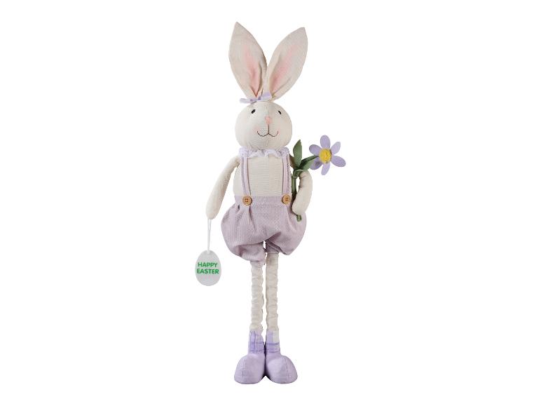 Plyšový velikonoční zajíček ve fialových lacláčích, držící květinu a cedulku 'Happy Easter'.