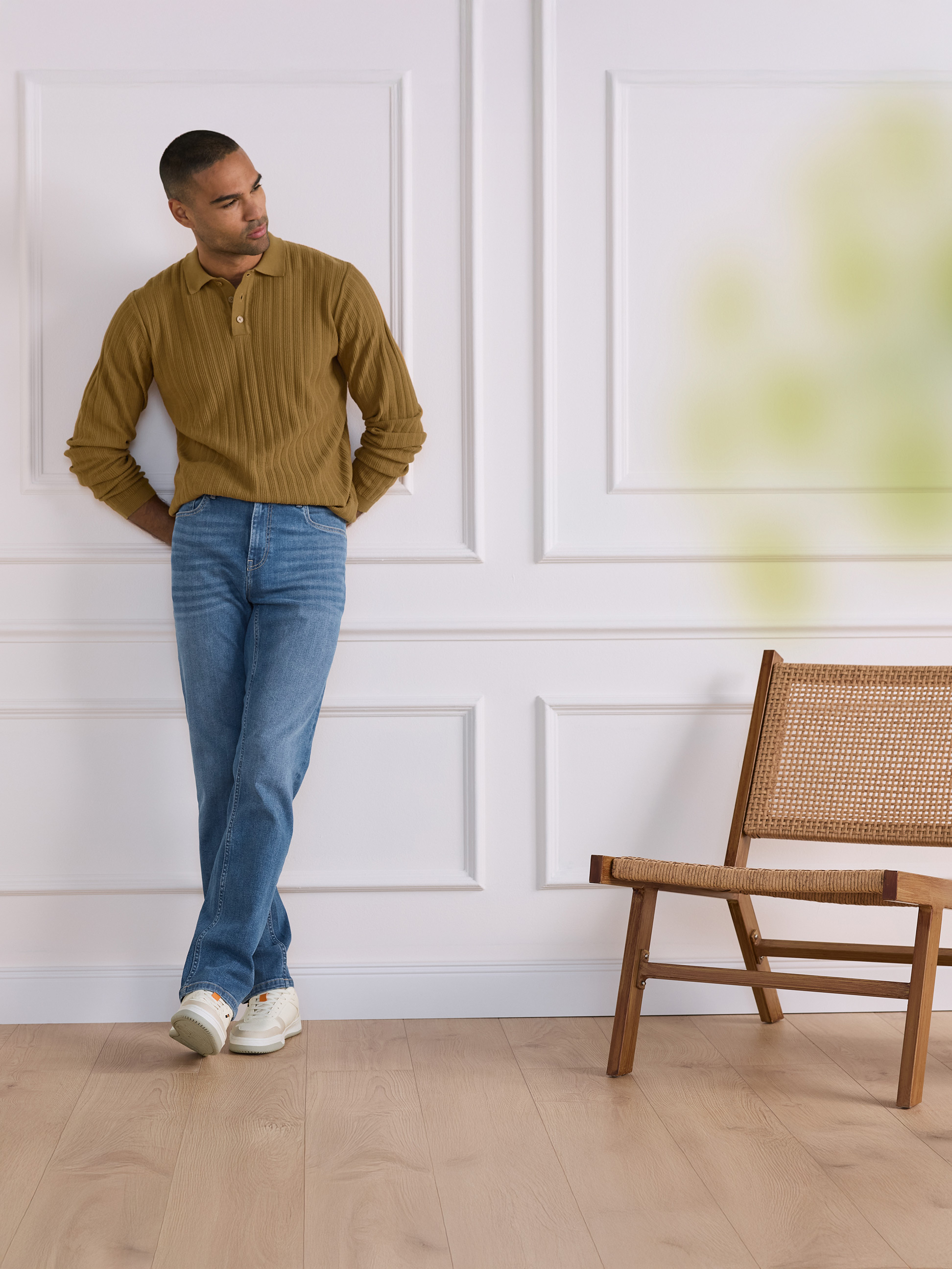 Muž v hnědém žebrovaném polotričku a džínách opřený o bílou zeď s dřevěnou židlí.