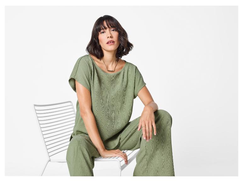 Žena v zeleném texturovaném topu a odpovídajících širokých kalhotách, sedící na bílé židli.