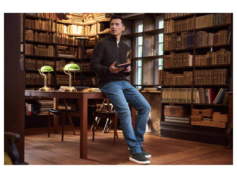 Muž v hnědém svetru a džínách sedí na stole v knihovně a drží knihu.