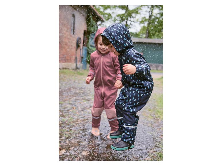 Dvě děti si hrají v kalužích v nepromokavých kombinézách a holínkách.