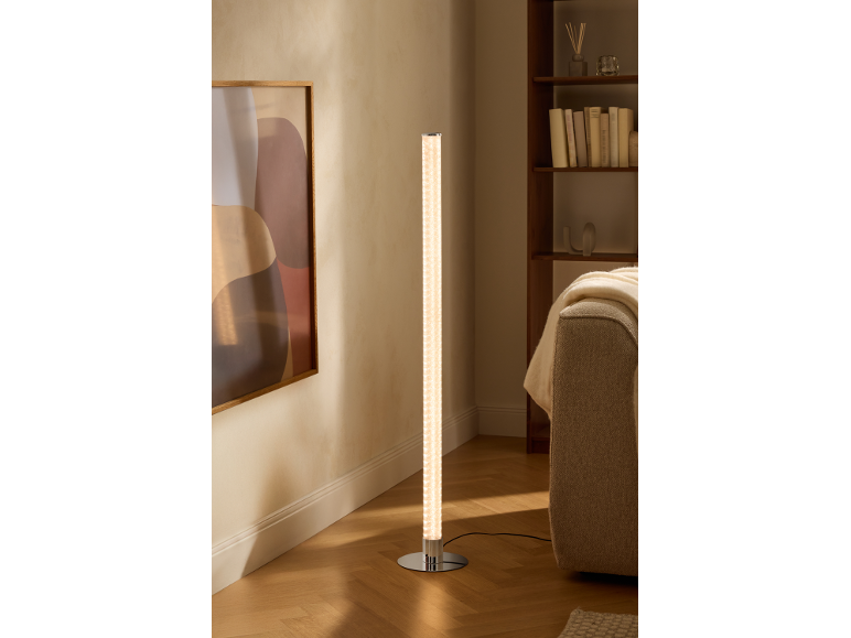 Válcová stojací lampa s chromovou základnou osvětluje moderní obývací pokoj s knihovnou a pohovkou.
