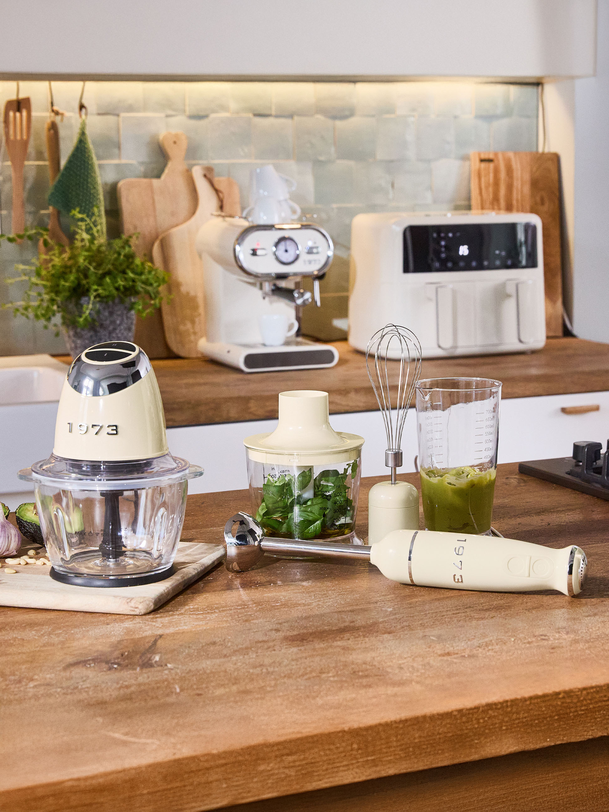 Krémové kuchyňské spotřebiče, včetně sekáčku, tyčového mixéru a kávovaru, na dřevěné desce.