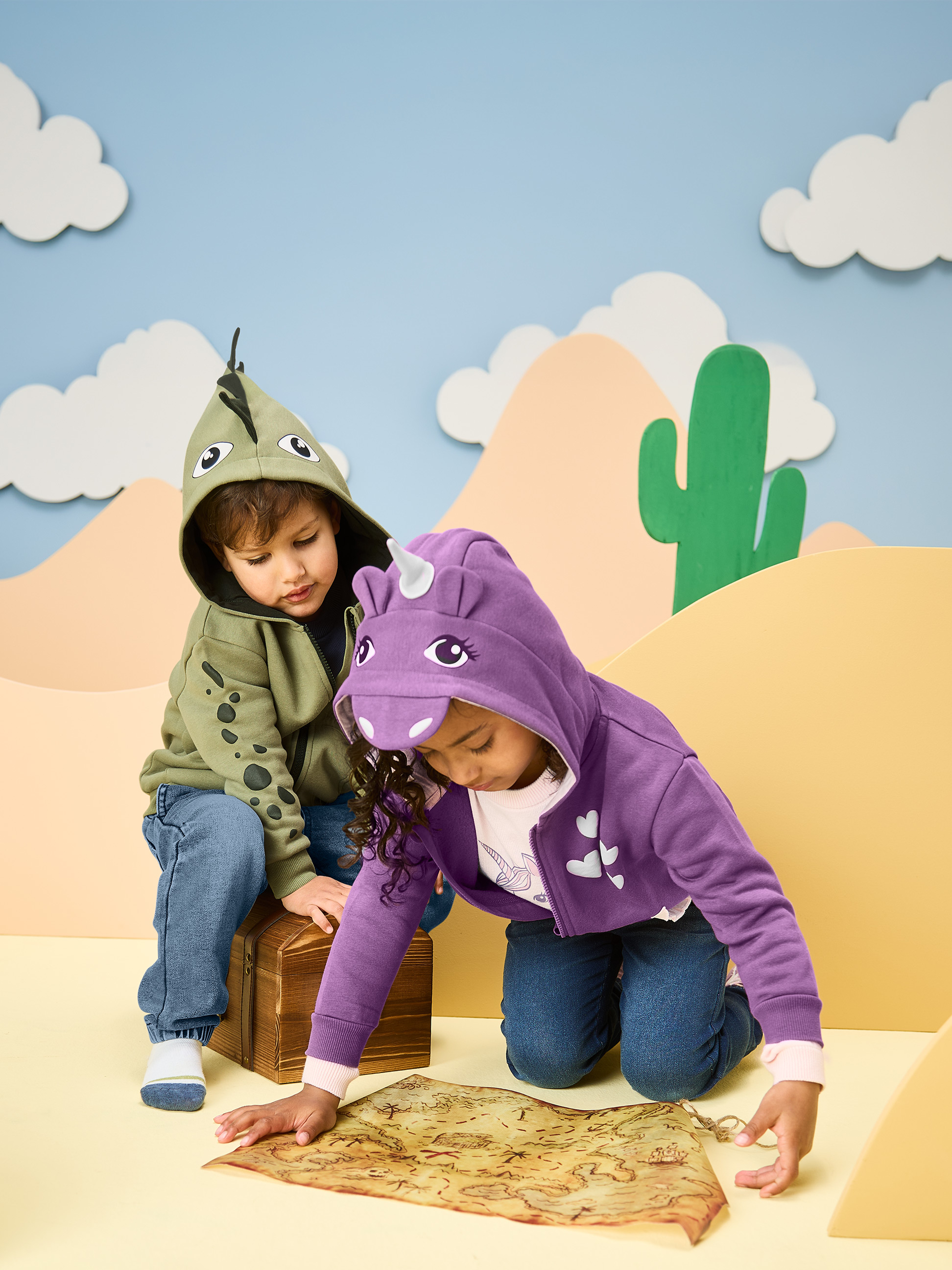 Děti v mikinách s kapucí dinosaura a jednorožce, prohlížející mapu pokladu.